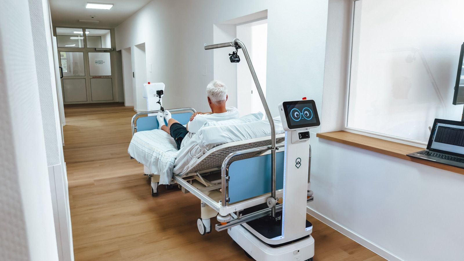 Transportroboter bewegt ein Krankenhausbett durch einen Stationsflur, im Bett liegt eine Person.